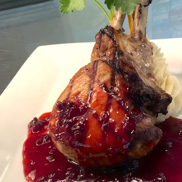 Grilled pork chop with a rich red berry sauce, mashed potatoes, and parsley garnish on a white plate.